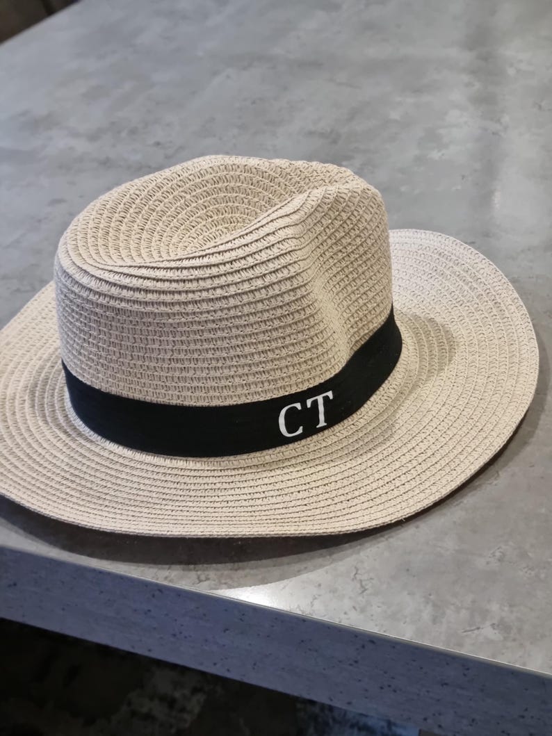 May include: A beige straw hat with a black band and the initials "CT" embroidered in white.
