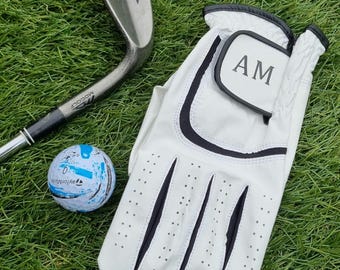 May include: White golf glove with black trim and a black patch with the initials "AM" on it. A golf ball and a golf club are also in the image.