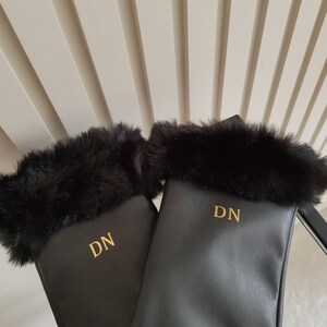 May include: A pair of black gloves featuring a soft black faux fur trim at the cuffs. Each glove is personalised with the gold-coloured embroidered initials DN centred on the back of the hand, providing a clear and distinct contrast against the black material.
