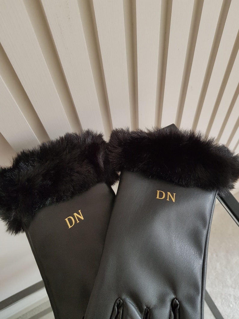 May include: A pair of black gloves featuring a soft black faux fur trim at the cuffs. Each glove is personalised with the gold-coloured embroidered initials DN centred on the back of the hand, providing a clear and distinct contrast against the black material.