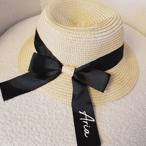 May include: A beige straw hat with a black ribbon and bow. The ribbon has the name "Aria" written on it in white.