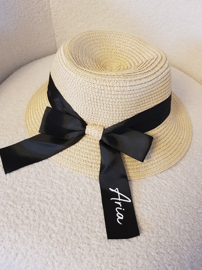 May include: A beige straw hat with a black ribbon and bow. The ribbon has the name "Aria" written on it in white.
