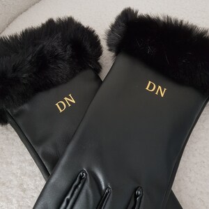 May include: A pair of black gloves featuring a smooth finish and a plush black faux fur trim at the cuffs. Each glove is personalised with the gold-coloured embroidered initials DN centred on the back of the hand, providing a clear contrast.
