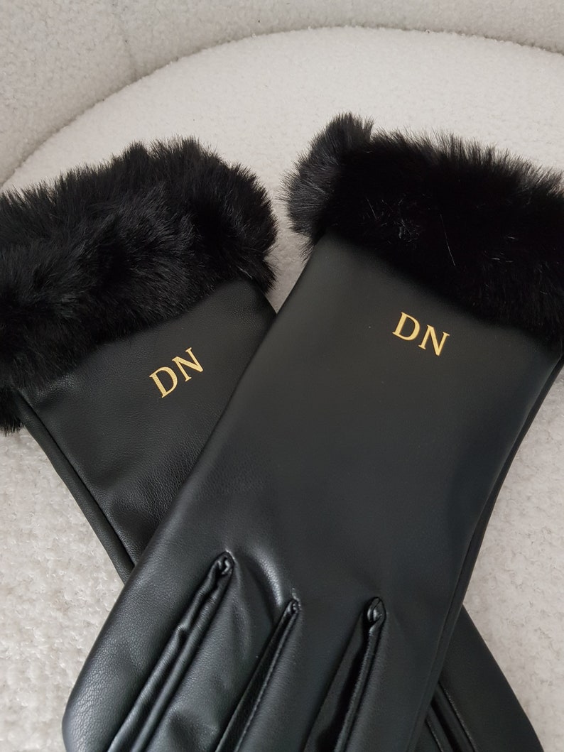 May include: A pair of black gloves featuring a smooth finish and a plush black faux fur trim at the cuffs. Each glove is personalised with the gold-coloured embroidered initials DN centred on the back of the hand, providing a clear contrast.