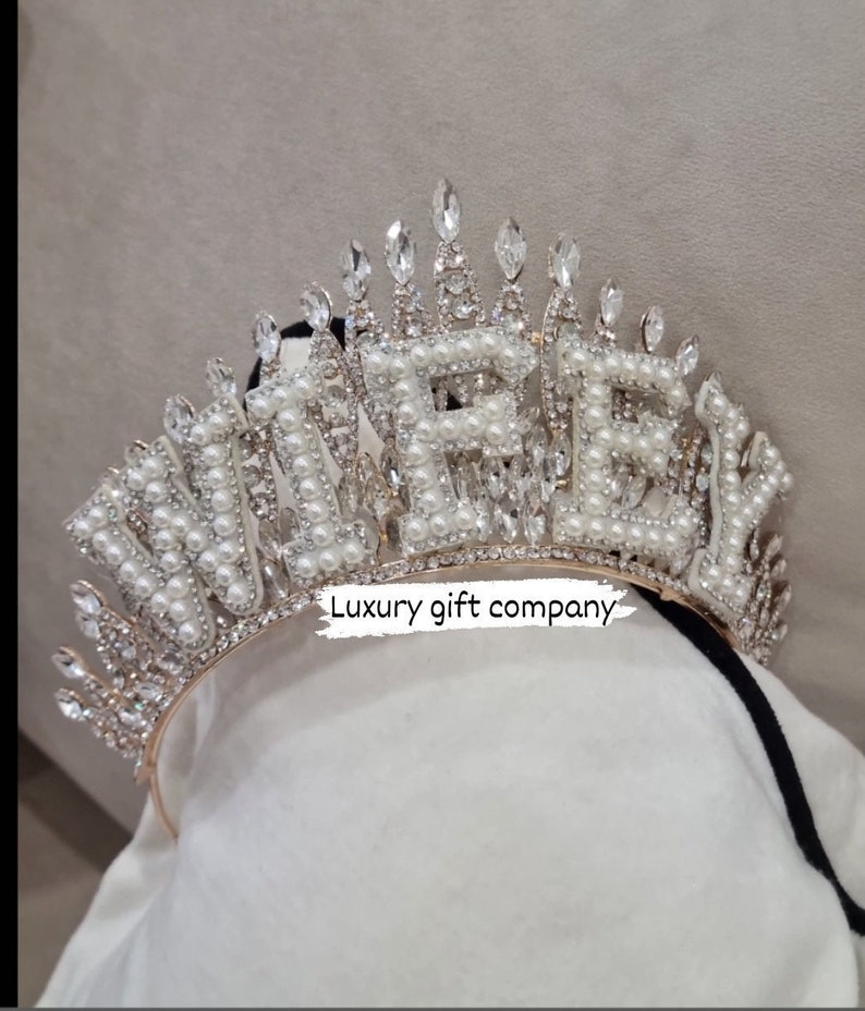 May include: A gold-tone headband with the word "WIFEY" spelled out in white pearls and rhinestones. The headband is decorated with a crown of smaller rhinestones.