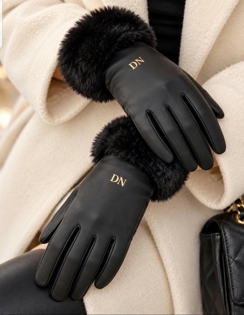 May include: A pair of black gloves featuring a smooth finish and plush black faux fur cuffs at the wrists. Each glove is embroidered with the gold-coloured letters DN on the back of the hand, positioned just below the fur trim for a distinct look.