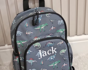 Könnte beinhalten: Ein grauer Rucksack mit einem Dinosaurier-Druck und dem Namen "Jack" auf der Vorderseite gestickt. Der Rucksack hat einen schwarzen Reißverschluss und schwarze Träger.