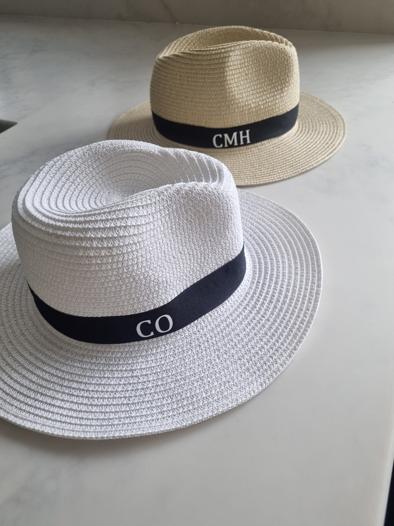 May include: Two straw hats with black bands. The hat on the left is white with the initials "CO" embroidered on the band. The hat on the right is beige with the initials "CMH" embroidered on the band.