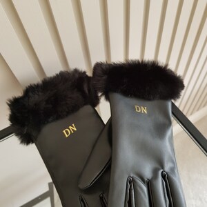 May include: A pair of black gloves featuring a soft black faux fur trim at the cuffs. Each glove is personalised with the gold embroidered initials DN centred on the back of the hand. The gloves have a smooth finish and a classic five-finger design.