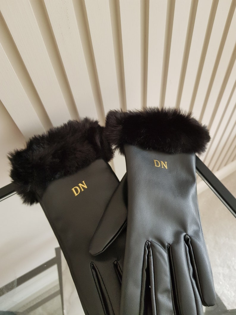 May include: A pair of black gloves featuring a soft black faux fur trim at the cuffs. Each glove is personalised with the gold embroidered initials DN centred on the back of the hand. The gloves have a smooth finish and a classic five-finger design.