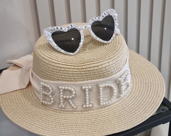 May include: A beige straw sun hat with a cream ribbon and the word "BRIDE" in pearl beads. Heart-shaped sunglasses with "BRIDE TO BE" are on top. The sunglasses are white with black lenses and rhinestone embellishments, perfect for a hen party.