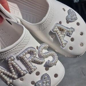 May include: A pair of white clogs with pearl embellishments spelling out "MRS" and "A" with a pearl heart on each shoe.
