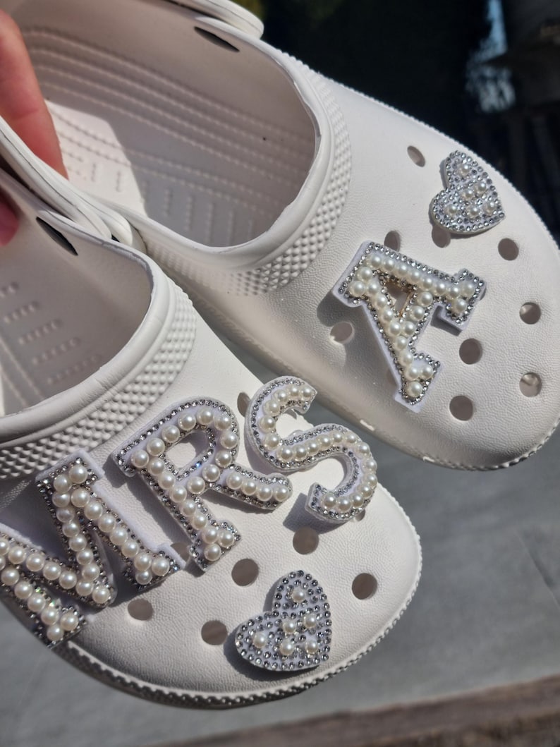 May include: A pair of white clogs with pearl embellishments spelling out "MRS" and "A" with a pearl heart on each shoe.