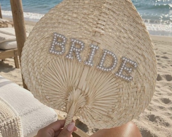 May include: A hand-held woven palm leaf fan with a teardrop shape. The word BRIDE is spelled out in large, capital letters across the centre of the fan, featuring a silver-toned border filled with small, white, round faux pearls.