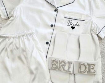 May include: White satin pyjama set with black trim and a pocket that says "Bride" with a heart. The set includes a short-sleeved top and shorts. The image also shows a pair of white slippers with "BRIDE" spelled out in rhinestones.