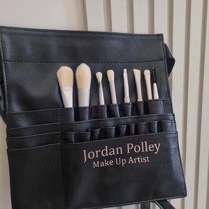 May include: Black leather makeup brush roll with a zipper closure and multiple pockets to hold brushes. The roll has the name "Jordan Polley Make Up Artist" printed on the front.