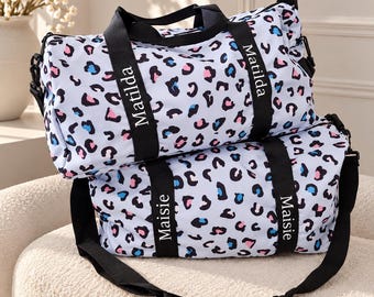 May include: Two duffel bags featuring a white base with a black, pink, and blue leopard print pattern. The top bag has black straps with the name Matilda printed in white, and the bottom bag has black straps with the name Maisie printed in white.