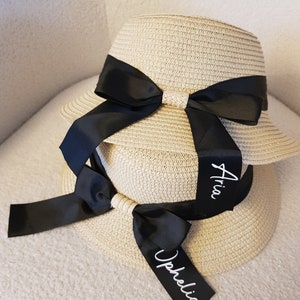 May include: Two straw sun hats with black ribbon bows and personalized names, "Aria" and "Ophelia", written in white on the ribbon.