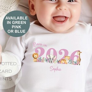 May include: White baby onesie with a pink "Born in 2026" design, featuring animal illustrations and the name "Sophie." The onesie also has text that says "Available in Green, Pink or Blue" and "Spotted Lizard Prints."