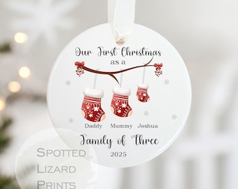 May include: A circular Christmas ornament with a white ribbon. It displays the text "Our First Christmas as a Family of Three", three hanging stockings, the names "Daddy, Mummy, Joshua" and the year 2025.