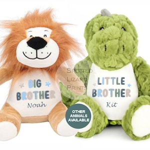Puede incluir: Un peluche de león color canela con melena marrón y un peluche de dinosaurio verde. El león tiene el texto BIG BROTHER Noah impreso en su vientre blanco. El dinosaurio tiene el texto LITTLE BROTHER Kit impreso en su vientre blanco. Ambos tienen estrellas.