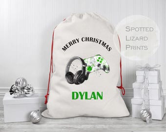 Puede incluir: Una bolsa de regalo de Navidad de color blanco roto con un cierre de cordón rojo. La bolsa presenta un gráfico de un mando de juegos y auriculares, con el texto "MERRY CHRISTMAS" y el nombre "DYLAN" en verde.