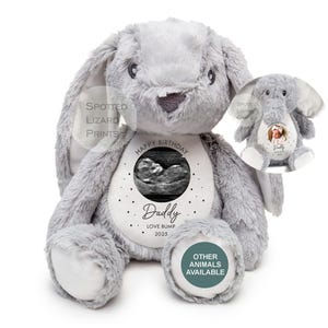May include: Gray plush bunny with a white circle on its chest. The circle has a black border and contains a black and white ultrasound image. The text "Happy Birthday Daddy Love Bump 2025" is printed on the circle. The bunny has a smaller white circle on its foot with the text "Other Animals Available" printed on it.