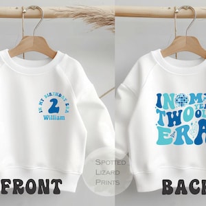 May include: White sweatshirt with blue text on the front that says "IN MY BIRTHDAY ERA" with the number "2" and the name "William". The back of the sweatshirt says "IN MY TWO YEAR OLD ERA" in blue.