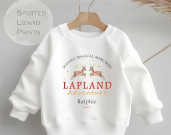 Może przedstawiać: Biała bluza z nadrukiem reniferów w kolorze czerwonym i złotym. Tekst głosi "Making Magical Memories Lapland Adventure Ralphie 2025".