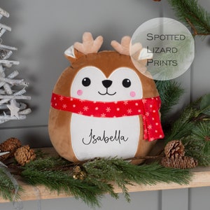 Puede incluir: Una almohada de reno de peluche marrón con una bufanda roja y copos de nieve. La almohada tiene una cara blanca con ojos negros y una nariz rosa. El nombre "Isabella" está bordado en la almohada.