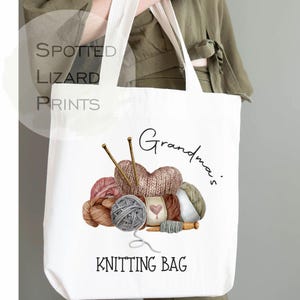 May include: White tote bag with a colorful knitting design. The bag features a heart-shaped knitted item, yarn balls, knitting needles, and the words "Grandma's Knitting Bag". The bag is held by a person.