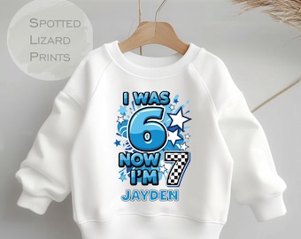 Könnte beinhalten: Weißes Sweatshirt mit dem Text "I WAS 6 NOW I'M 7 JAYDEN" in Blau und Schwarz. Die Zahl 6 ist blau mit weißem Rand, und die Zahl 7 ist ein schwarz-weißes Schachbrettmuster. Das Sweatshirt hat einen Rundhalsausschnitt und lange Ärmel.