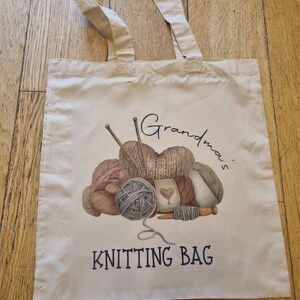 May include: Cream-colored tote bag with beige handles. The bag features a watercolor illustration of yarn balls, knitting needles, and the text "Grandma's KNITTING BAG" in navy blue. Perfect for carrying knitting supplies.
