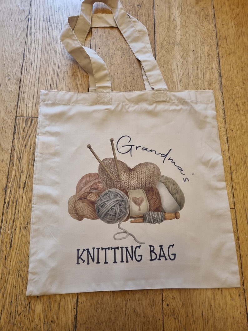 May include: Cream-colored tote bag with beige handles. The bag features a watercolor illustration of yarn balls, knitting needles, and the text "Grandma's KNITTING BAG" in navy blue. Perfect for carrying knitting supplies.