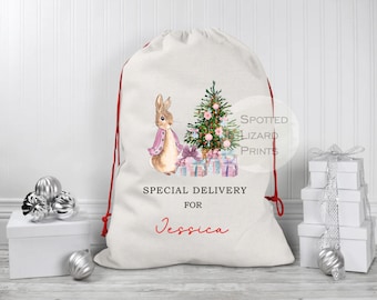 Puede incluir: Un saco de regalo navideño blanco con cierre de cordón rojo. El saco presenta una ilustración de acuarela de un conejo, un árbol de Navidad y regalos. El texto en el saco dice "SPECIAL DELIVERY FOR Jessica".