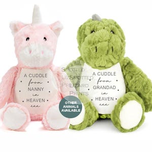 Könnte beinhalten: Ein rosa Plüscheinhorn und ein grüner Plüschdinosaurier, beide mit einem weißen Bauchfleck. Der Aufnäher des Einhorns trägt die Aufschrift A CUDDLE from NANNY in HEAVEN xxx. Der Aufnäher des Dinosauriers zeigt A CUDDLE from GRANDAD in HEAVEN xxx.