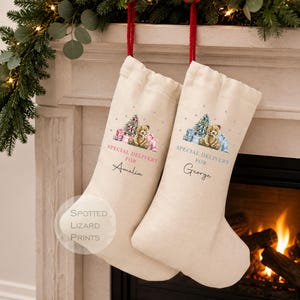 Puede incluir: Dos calcetines navideños de tela color hueso con lazos rojos para colgar. Cada uno presenta una ilustración en acuarela de un oso de peluche con regalos y un árbol, con el texto "SPECIAL DELIVERY FOR" seguido de los nombres "Amelia" y "George".