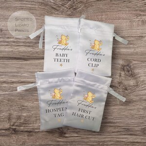 May include: Four white satin drawstring bags with Winnie the Pooh illustrations and personalized text. The bags are labeled "Freddie's Baby Teeth", "Freddie's Cord Clip", "Freddie's Hospital Tag", and "Freddie's First Hair Cut".