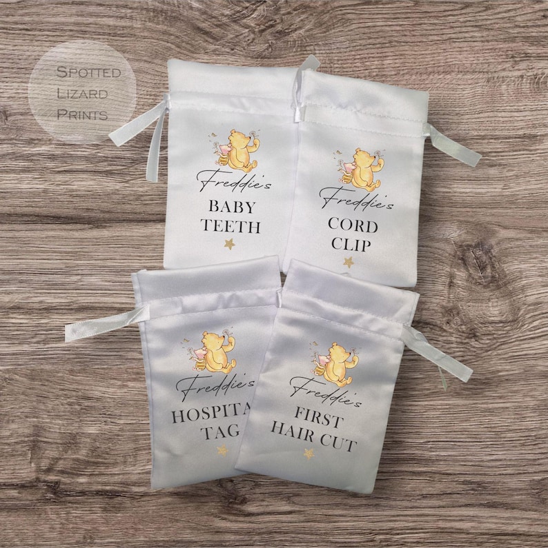 May include: Four white satin drawstring bags with Winnie the Pooh illustrations and personalized text. The bags are labeled "Freddie's Baby Teeth", "Freddie's Cord Clip", "Freddie's Hospital Tag", and "Freddie's First Hair Cut".