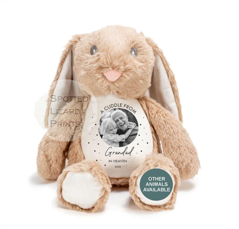 May include: A plush tan rabbit toy with long ears and a soft texture. The belly features a circular white patch with a black and white photograph of an adult and child, the text A CUDDLE FROM Grandad IN HEAVEN xxx, and small black dots.