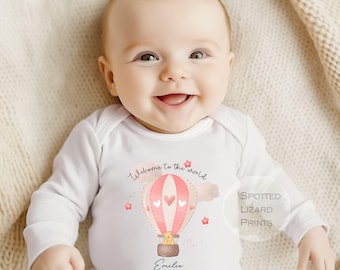 Könnte beinhalten: Ein weißer Langarmbody mit einem rosa-weißen Heißluftballon-Motiv. Der Ballon hat einen kleinen Bären im Inneren und den Text "Willkommen in der Welt" darüber. Der Body ist mit dem Namen "Emilie" personalisiert.