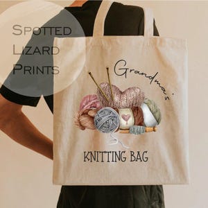 May include: Beige tote bag with a watercolor illustration of yarn balls, knitting needles, and a knitted heart. The words "Grandma's Knitting Bag" are printed on the bag. The bag is carried by a person.