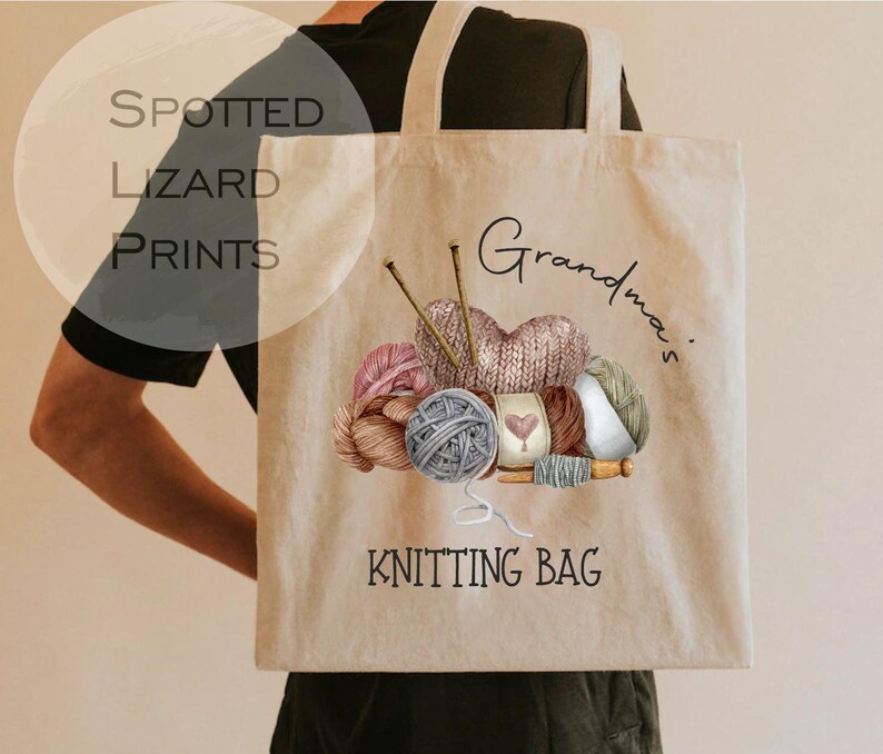 May include: Beige tote bag with a watercolor illustration of yarn balls, knitting needles, and a knitted heart. The words "Grandma's Knitting Bag" are printed on the bag. The bag is carried by a person.