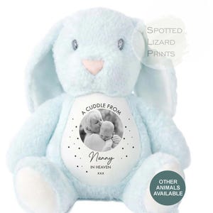 May include: A light blue plush rabbit toy with a white circular patch on its chest. The patch features a black and white photograph of an adult and a baby, with the text A CUDDLE FROM Nanny IN HEAVEN xxx and small star icons surrounding the image.