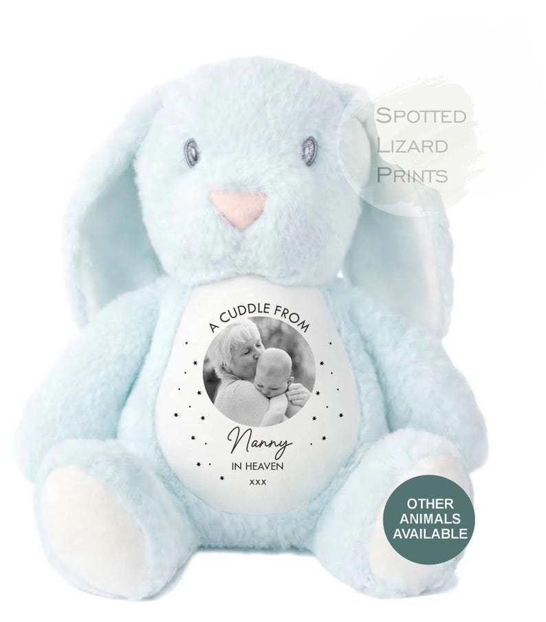 May include: A light blue plush rabbit toy with a white circular patch on its chest. The patch features a black and white photograph of an adult and a baby, with the text A CUDDLE FROM Nanny IN HEAVEN xxx and small star icons surrounding the image.
