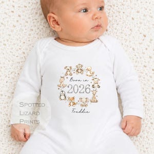 Op de afbeelding: Een wit babyrompertje met lange mouwen voorzien van een cirkelvormige krans van aquarel-stijl babydieren rondom de tekst Born in 2026 en de naam Freddie. De dieren zijn onder andere een hond, aap, eend, paard, pinguïn, vos, olifant en giraf.