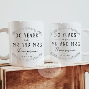 Op de afbeelding: Twee witte keramische mokken met de tekst "30 YEARS as MR AND MRS Thompson 15.05.1995" in zwart schrift. Elke mok heeft een decoratief kransontwerp. De mokken worden gepresenteerd op een houten krat.