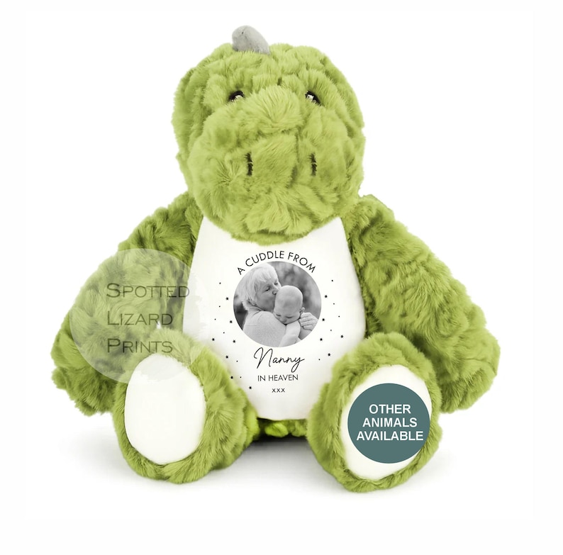 May include: A plush green dinosaur toy with a white belly patch featuring a circular black and white photograph of an adult holding a baby. The text reads A CUDDLE FROM Nanny IN HEAVEN XXX. The toy has soft textured fur and white foot pads.