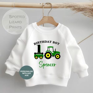 May include: White sweatshirt featuring a green and yellow tractor design, the words "BIRTHDAY BOY" and "Spencer". The sweatshirt also has the text "PERSONALISE WITH ANY AGE".