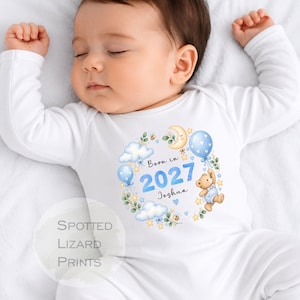 Puede incluir: Un body blanco de manga larga para bebé con un estampado gráfico de un osito de peluche, globos azules, nubes, estrellas y abejas. El texto dice Born in 2027 y Joshua. Una marca de agua circular en la esquina muestra el texto Spotted Lizard Prints.
